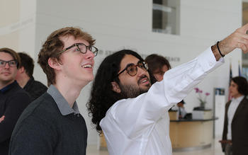 A young man in glasses looks at what a young man in a white shirt is pointing at.